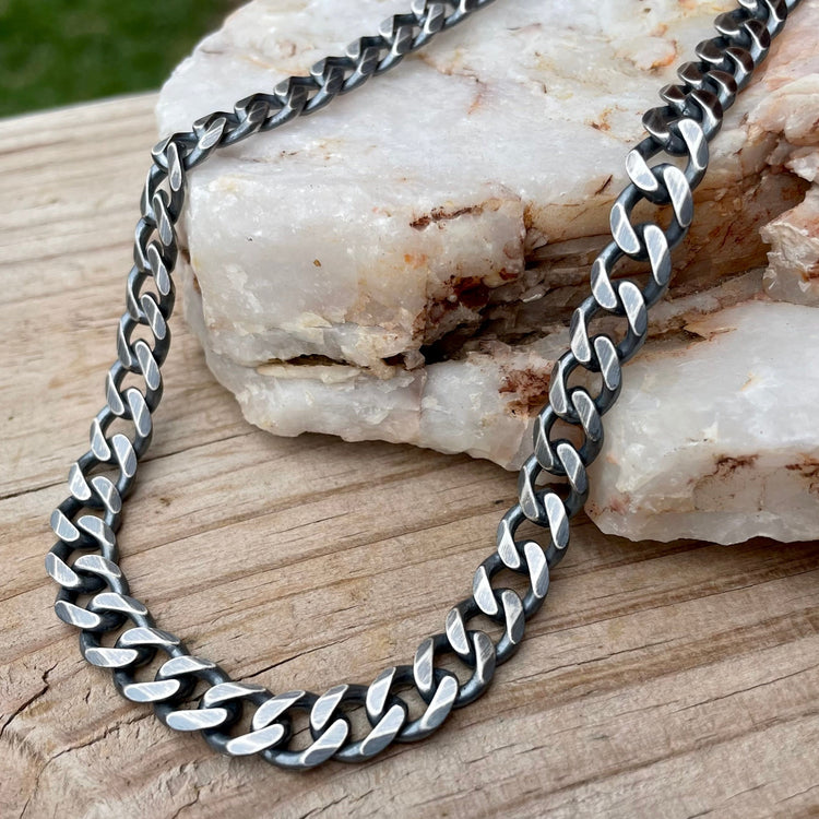 Heavy oxidized sterling silver curb chain necklace 10mm in width with lobster clasp, displayed on stone and wood. 