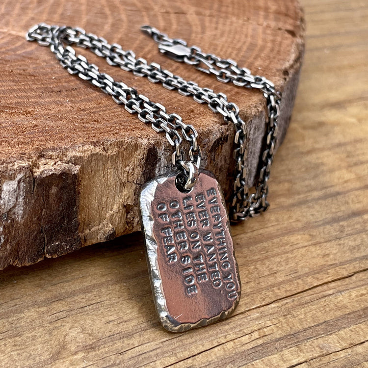 Personalized, custom engraved one inch sterling silver pendant shown with 2.7mm oxidized 925 silver cable chain. Hand forged with distressed and textured edges with deep hand struck text. This pendant reads "everything you ever wanted lies on the other side of fear". Shown on wood back ground. 