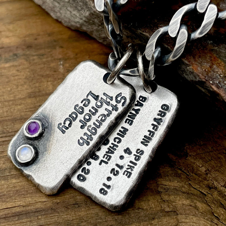 2 large sterling silver pendants with custom engraved words of strength, honor, legacy as well as names and birthdates of children. Shown with two birthstones hanging from a 9mm oxidized sterling silver curb chain displayed on wood. 