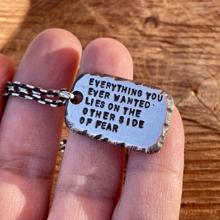 Personalized, custom engraved one inch sterling silver pendant shown with 2.7mm oxidized 925 silver cable chain. Hand forged with distressed and textured edges with deep hand struck text. This pendant reads "everything you ever wanted lies on the other side of fear". Shown in hand. 