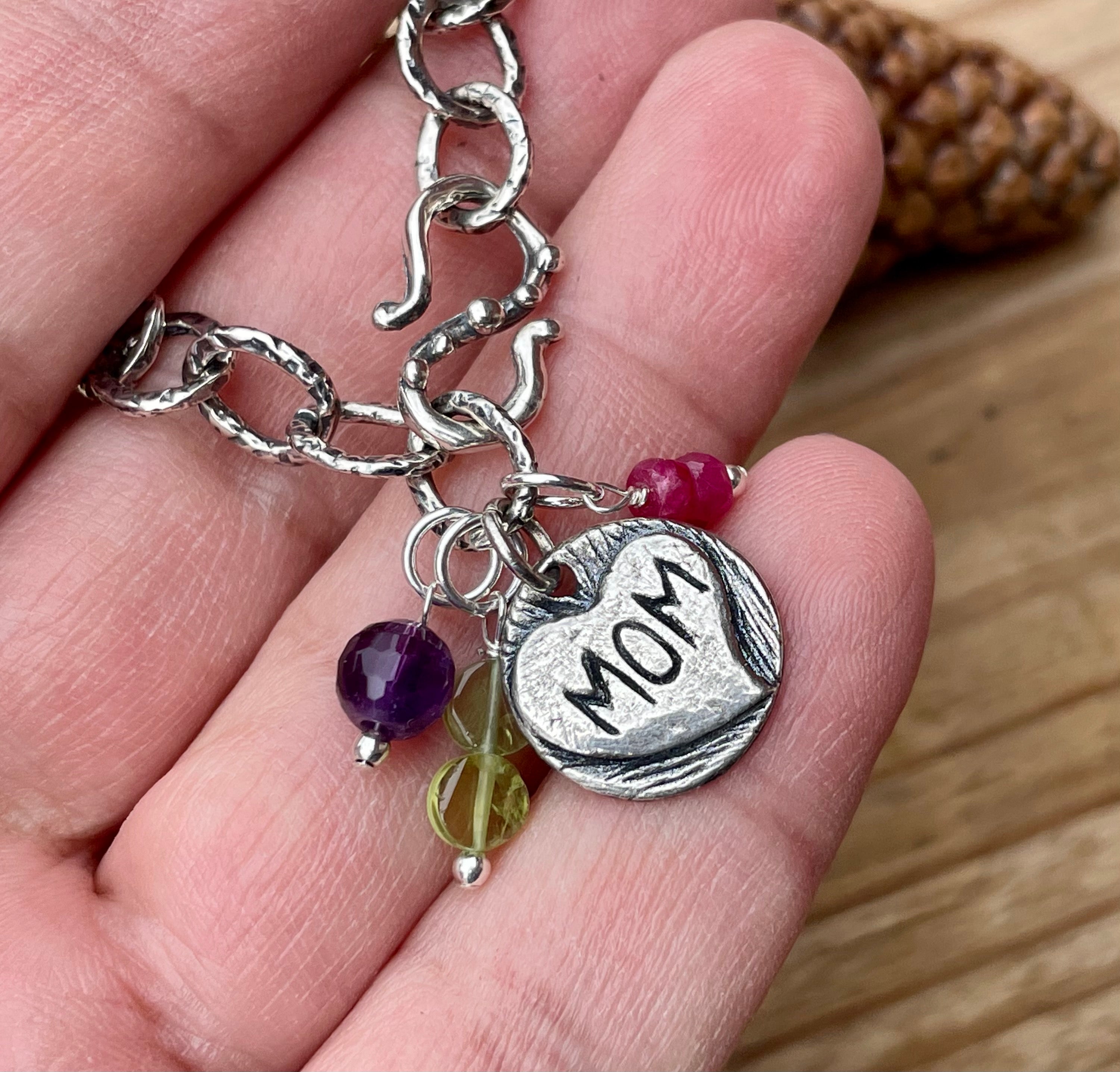 Artisan sterling silver charm bracelet with decorative s hook clasp and charm that says Mom inside of heart. Shown with three birthstones in hand. 