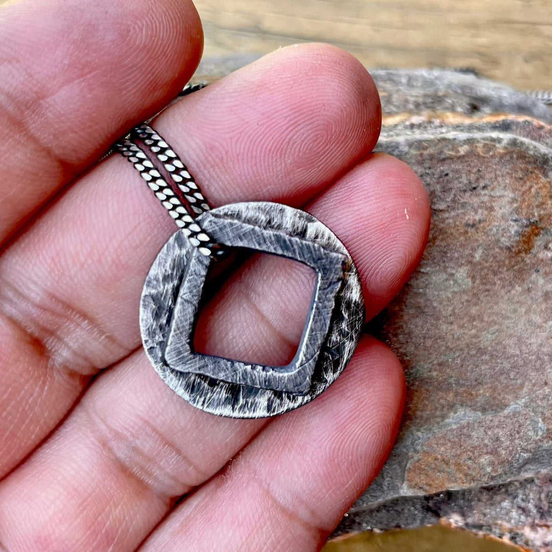 Hand carved pendants stacked together hanging from 2mm curb chain. Bottom pendant is an oxidized circle and top pendant is an oxidized square pendant. Free floating charms on 2mm curb chain. Shown in hand.