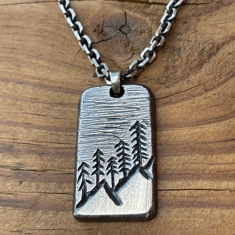 Oxidized sterling silver hand carved rectangle pendant with mountain and tree scene. Shown with 4mm cable chain. Back side can be personalized. Displayed on wood background. 