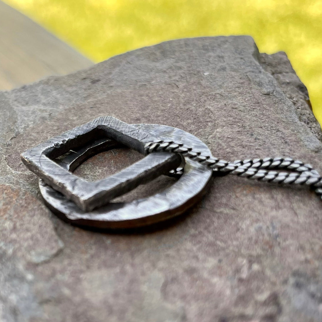 Side view of Hand carved pendants stacked together hanging from 2mm curb chain. Bottom pendant is an oxidized circle and top pendant is an oxidized square pendant. Free floating charms on 2mm curb chain. Shown on stone background. 