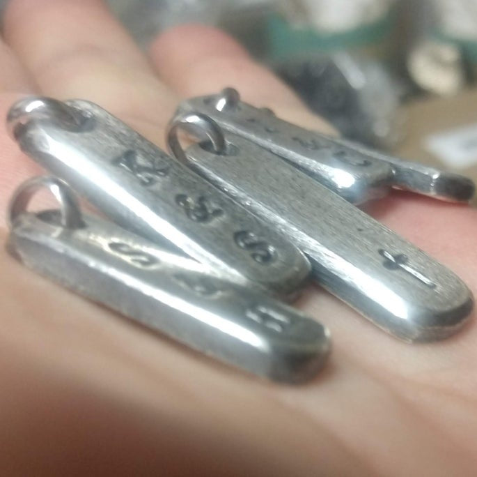 Five Sterling Silver initial bar tags shown with faint letters and a cross in side view, held in hand. 