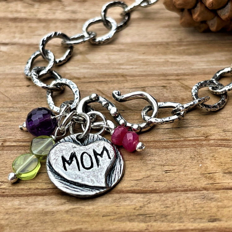 A silver bracelet with a heart-shaped charm labeled 'Mom', surrounded by small colorful gemstone charms, on a wooden background. Chain links are textured and hammered and clasp is a decorative hook. 